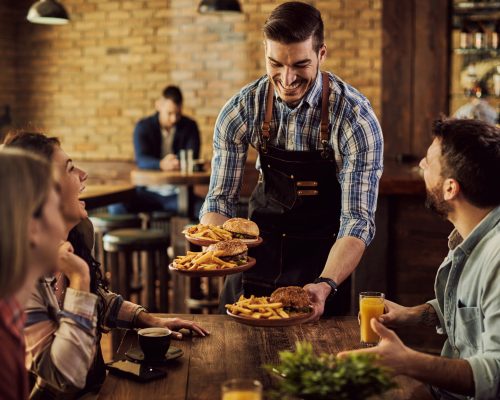 Group of happy friends having fun while waiter is serving them food in a pub. Focus is on waiter.